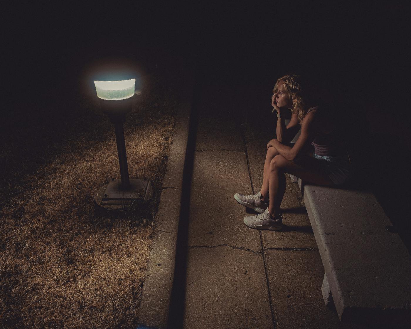 Night, a woman alone sits on a stone bench in front of a pathway light pondering life