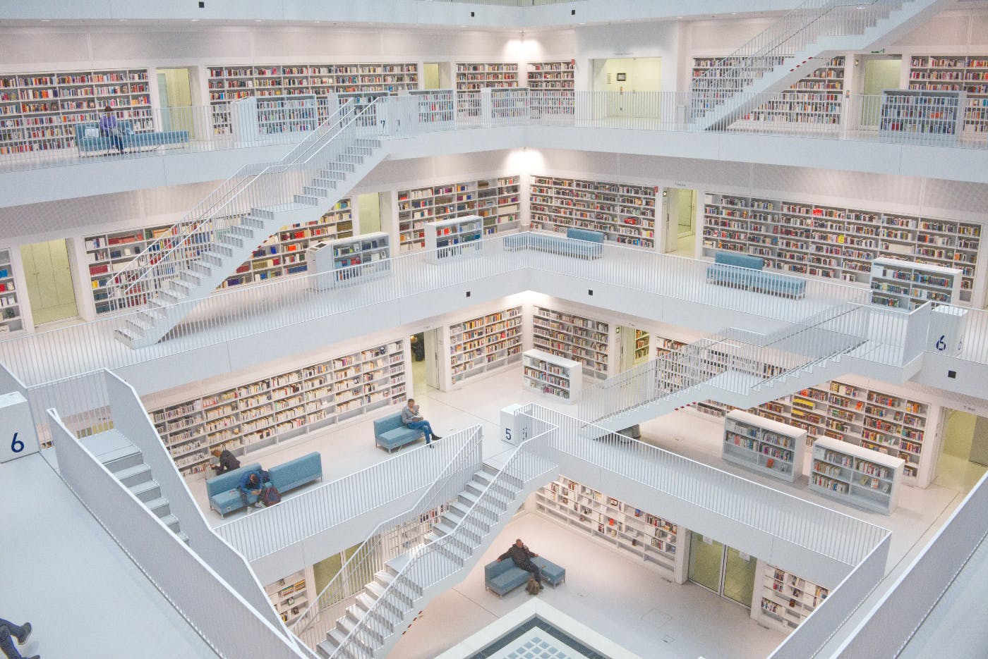 A library with white walls and stairs People sitting on benches reading.