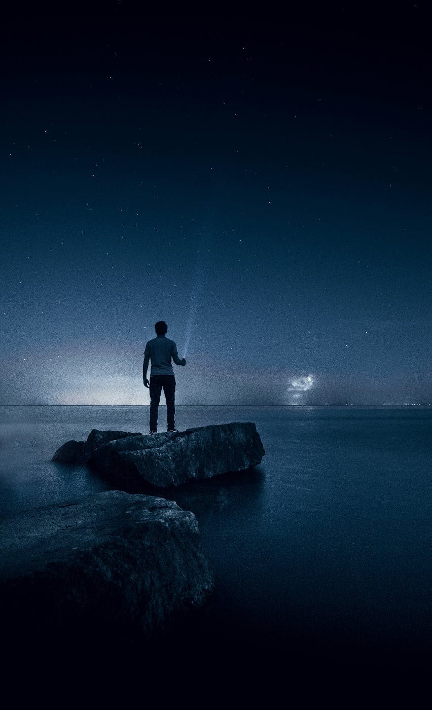 A man standing on a rock in water aiming a flashlight up into a star filled night sky