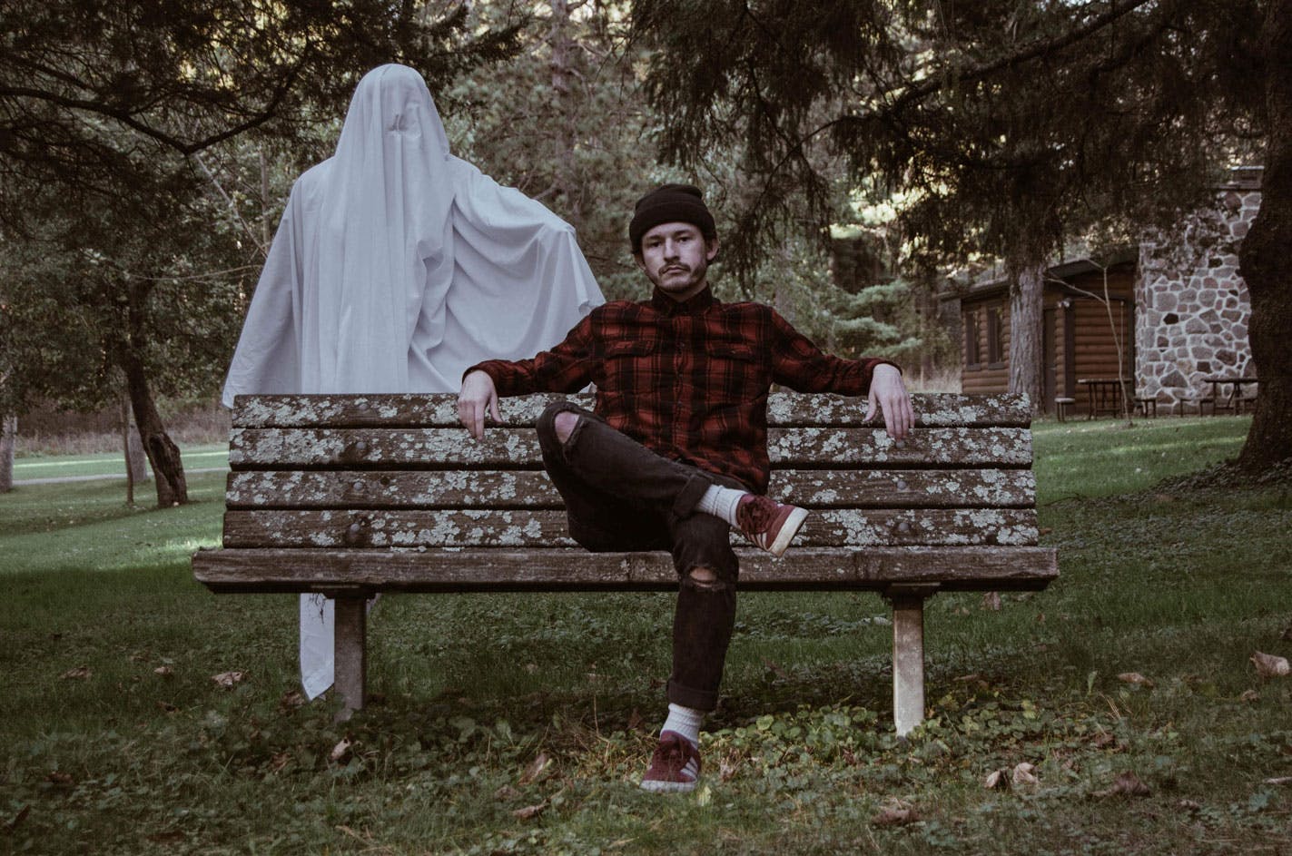 hipster man sitting on a bench in a red plaid shirt with a sheet ghost lingering behind him