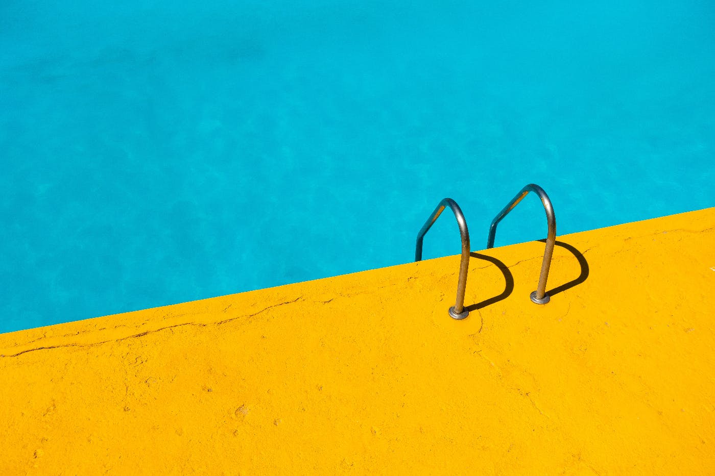 Blue water of a swimming pool, the yellow ocra deck and the handles of a stainless steel ladder