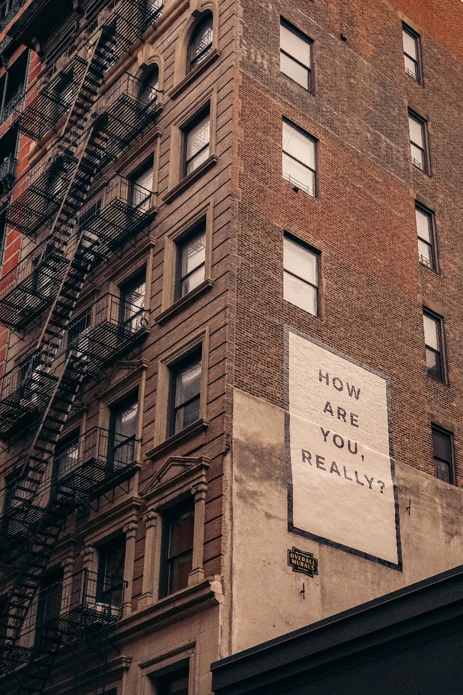 A brick apartment building with fire escapes and a sign on the side reading: How Are You Really?