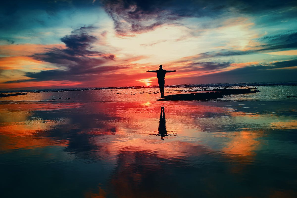 A person stanidng on wet sand witht he colors of the sunset reflected on the water, land and sky meld.