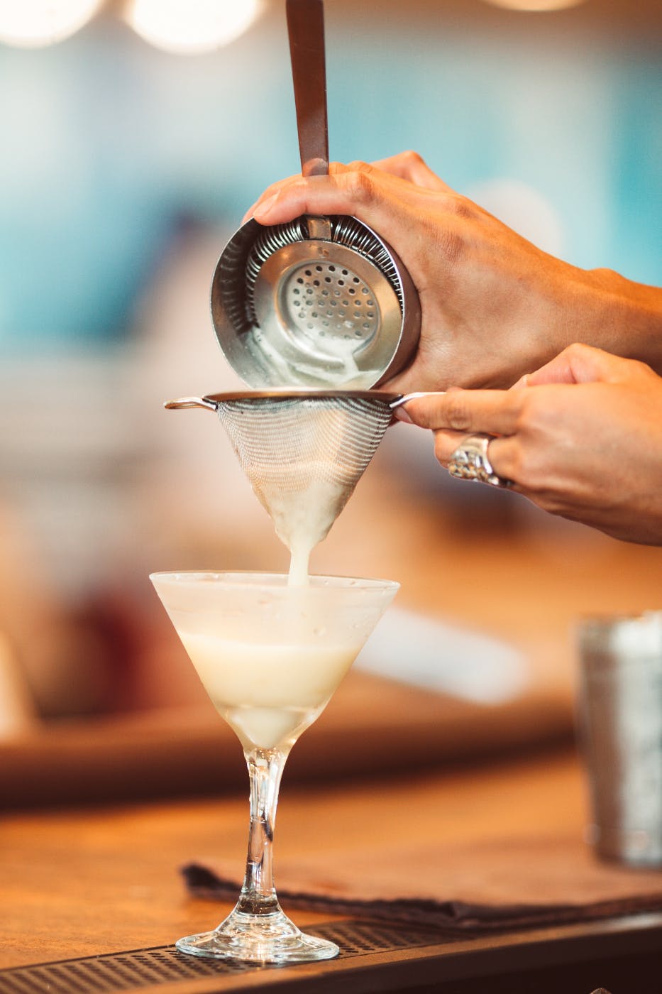 A bar tender pouring a white cocktail from a shaker to a strainer into a glass