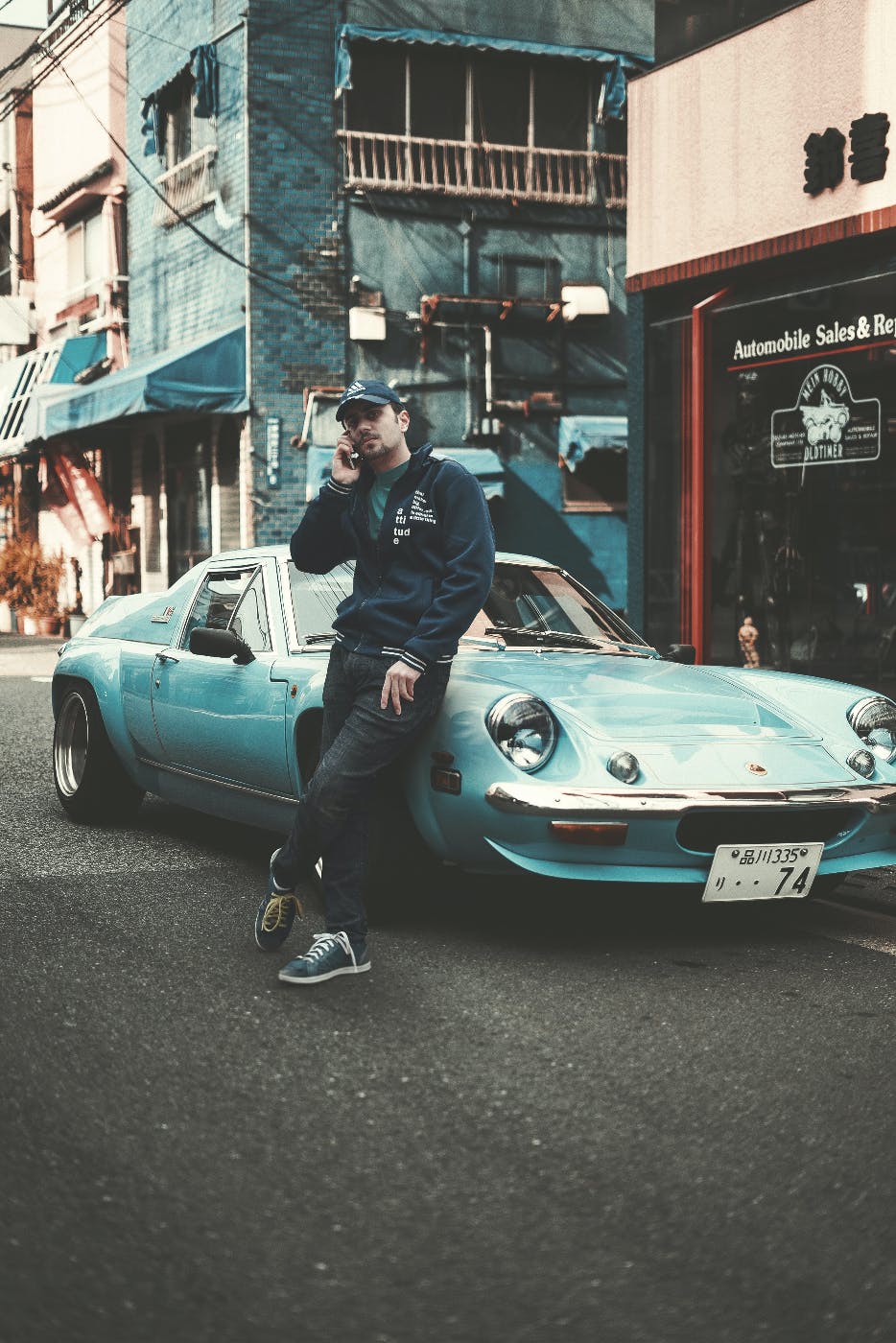 A guy leaning against a beat up blue sports car, talking on his smartphone