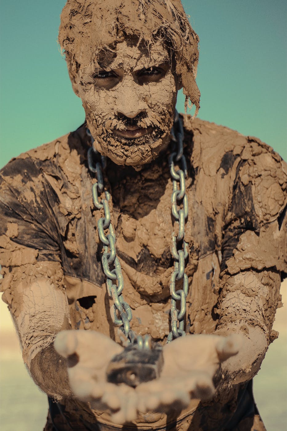 A man covered in mud with a chain around his neck holding the lock to the chain in his two outstretched hands