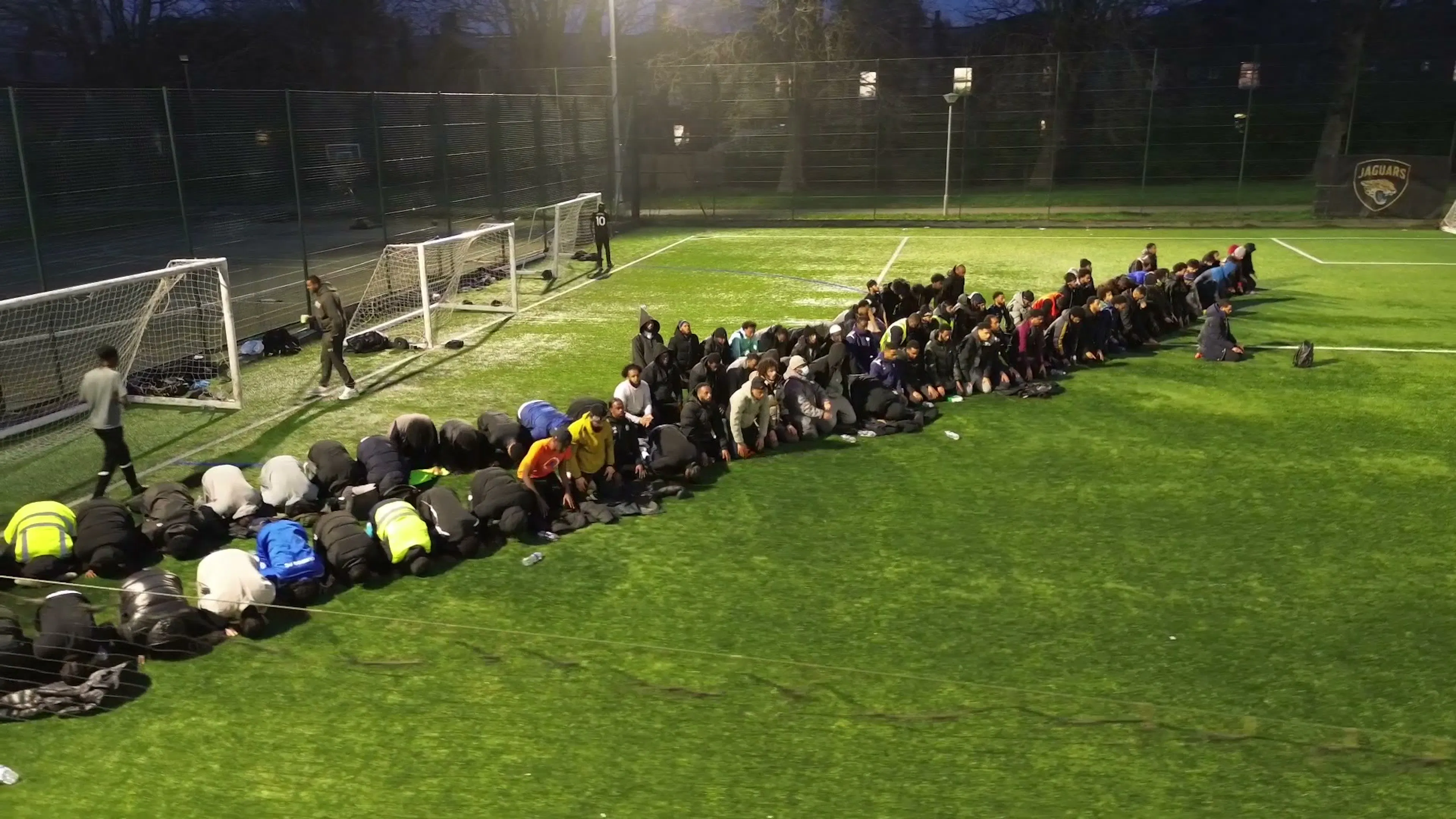 Players and spectators praying together in a line across a floodlit pitch