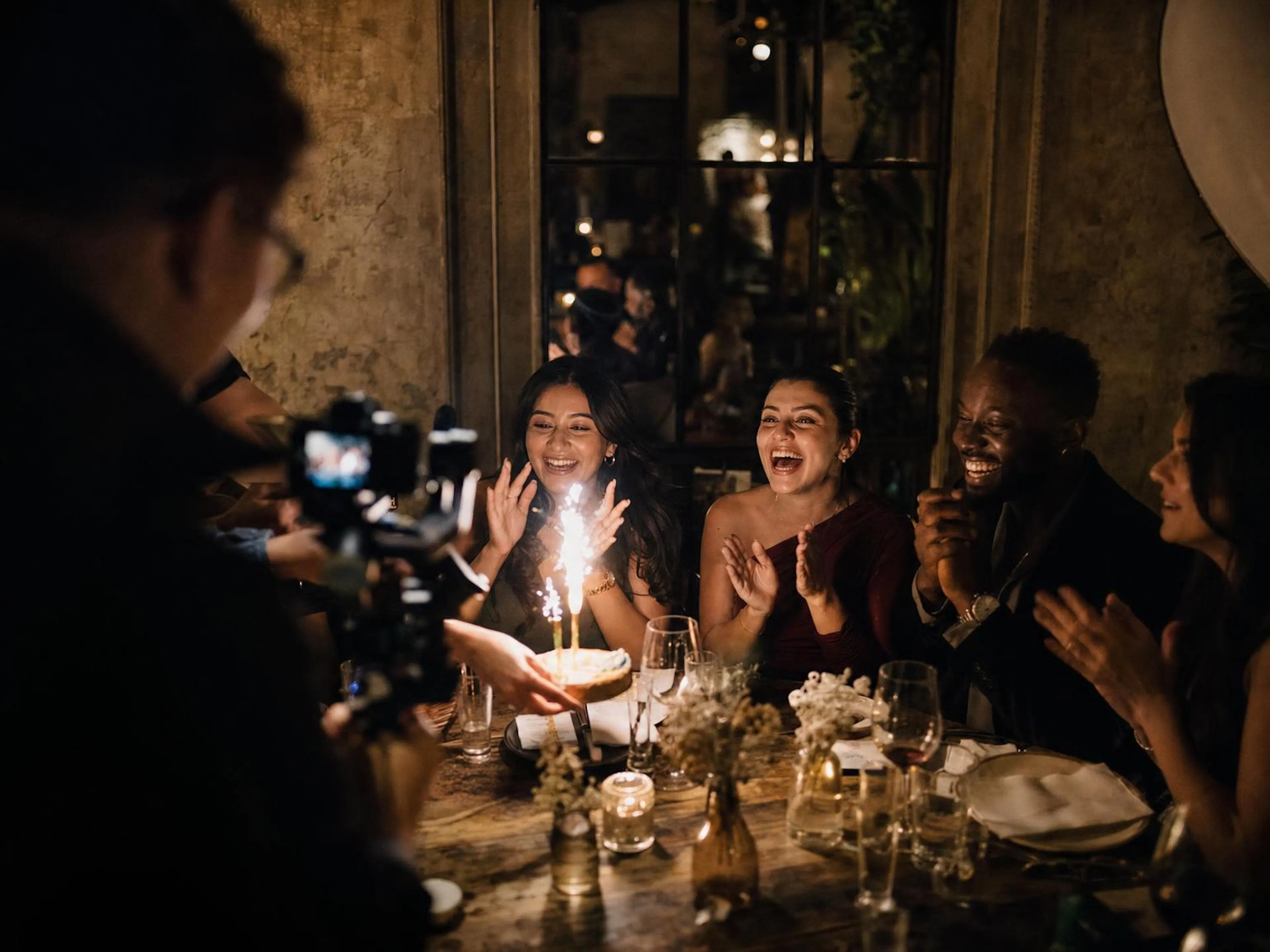 Friends cheering as a birthday cake with sparklers arrives, a videographer filming