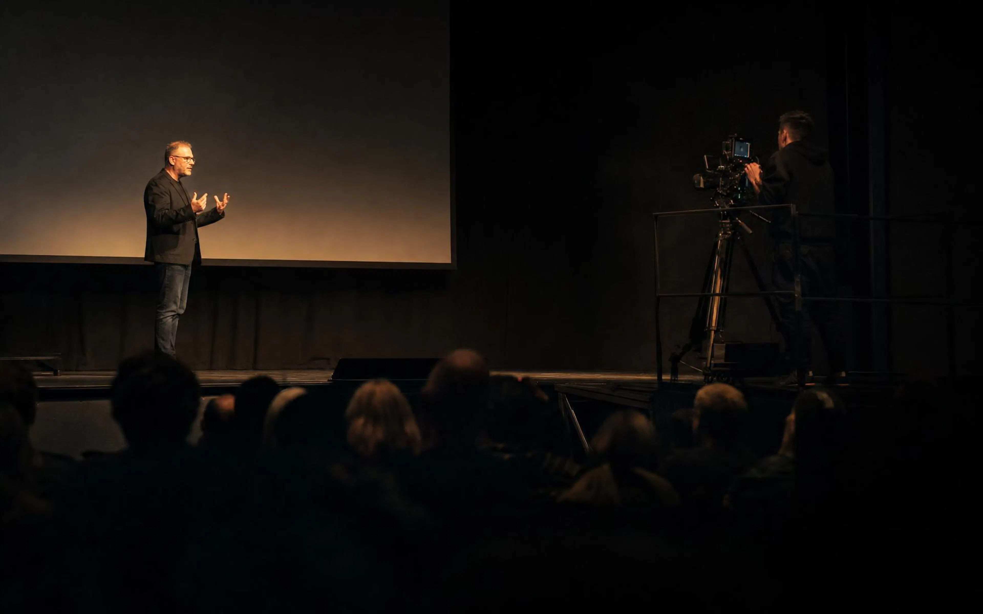 A speaker on a dark conference stage under a single spotlight, filmed from the audience