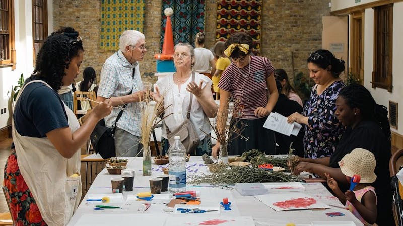 A group of people are gathered around a table covered with paints, prints and plants.