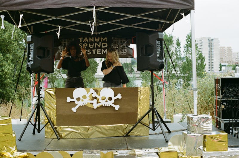 A DJ booth at a summer festival in the foreground, greenery in the background. Two people behind the DJ deck have their hands up, wearing black T-shirts. The booth is under a tent, with 2 speakers standing to the decks' sides. The deck is decorated with skulls, in line with a pirate theme. There is some audio equipment and boxes around the booth. 