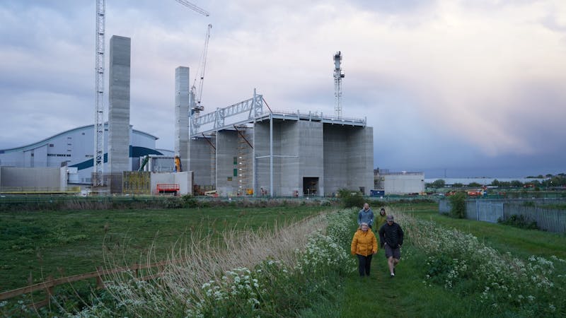 The image shows a group of people walking on a grassy path next to a construction site with large concrete buildings and cranes. The sky is cloudy with some light breaking through. The people are wearing casual outdoor clothing, suggesting a cool day.