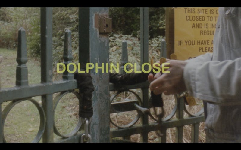 Film still showing the title "Dolphin Close" in the middle in yellow. Someone's hand are seen opening a lock on a metal, greenish gate, on which there is a yellow notice board. Behind the gate, greenery, grass and trees can be seen. 