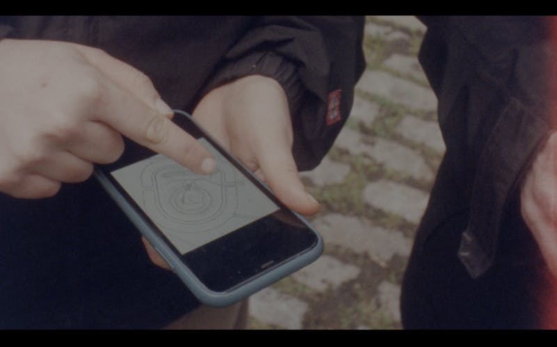 The film still shows a close up of a person's hands holding a smartphone and pointing to a map on their phone. The other person's silhouette is visible a little to the right. The photo is from an analog camera, colours are muted. 