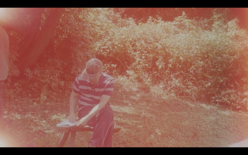 Analog film still showing a man sitting on a small bench, looking at a piece of paper on the bench. He is dressed in long trousers and striped T-shirt. He is sitting in a natural environment, there are trees and bushes behind him, grass on the ground. The still has a reddish filter applied, distorting natural colours and giving it an older feel. 