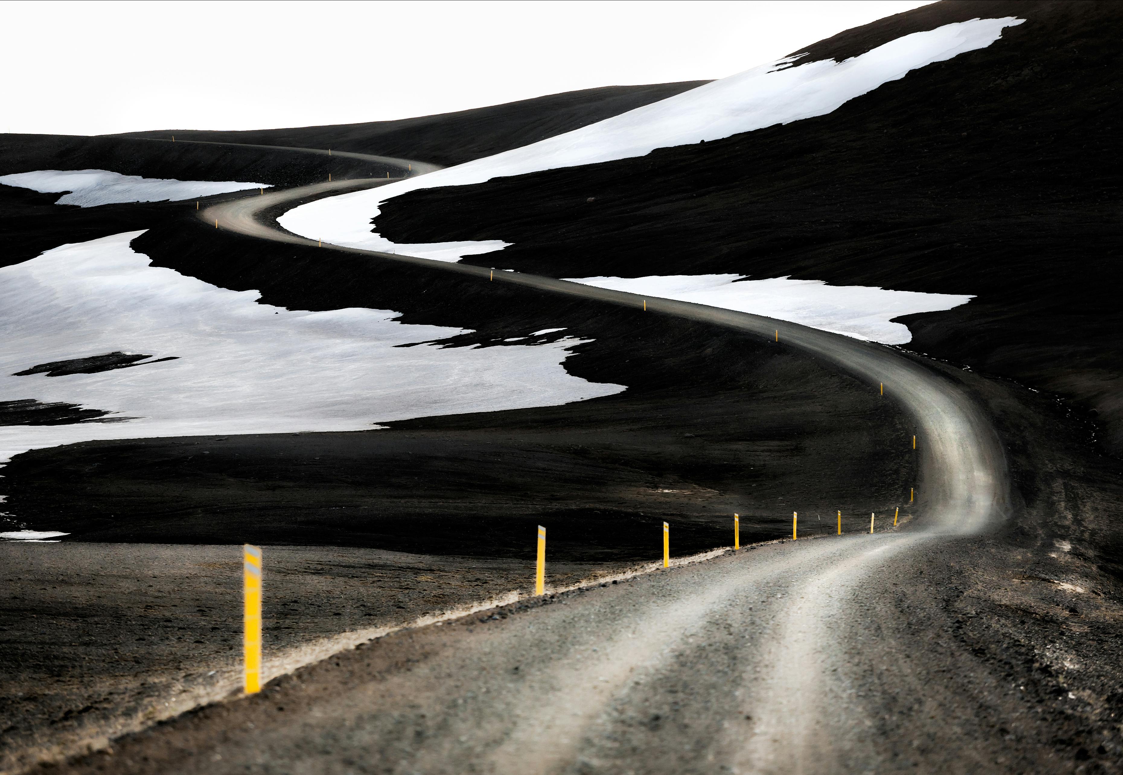 Winter road through black sand, surrounded by snow-covered mountains and rugged landscape.