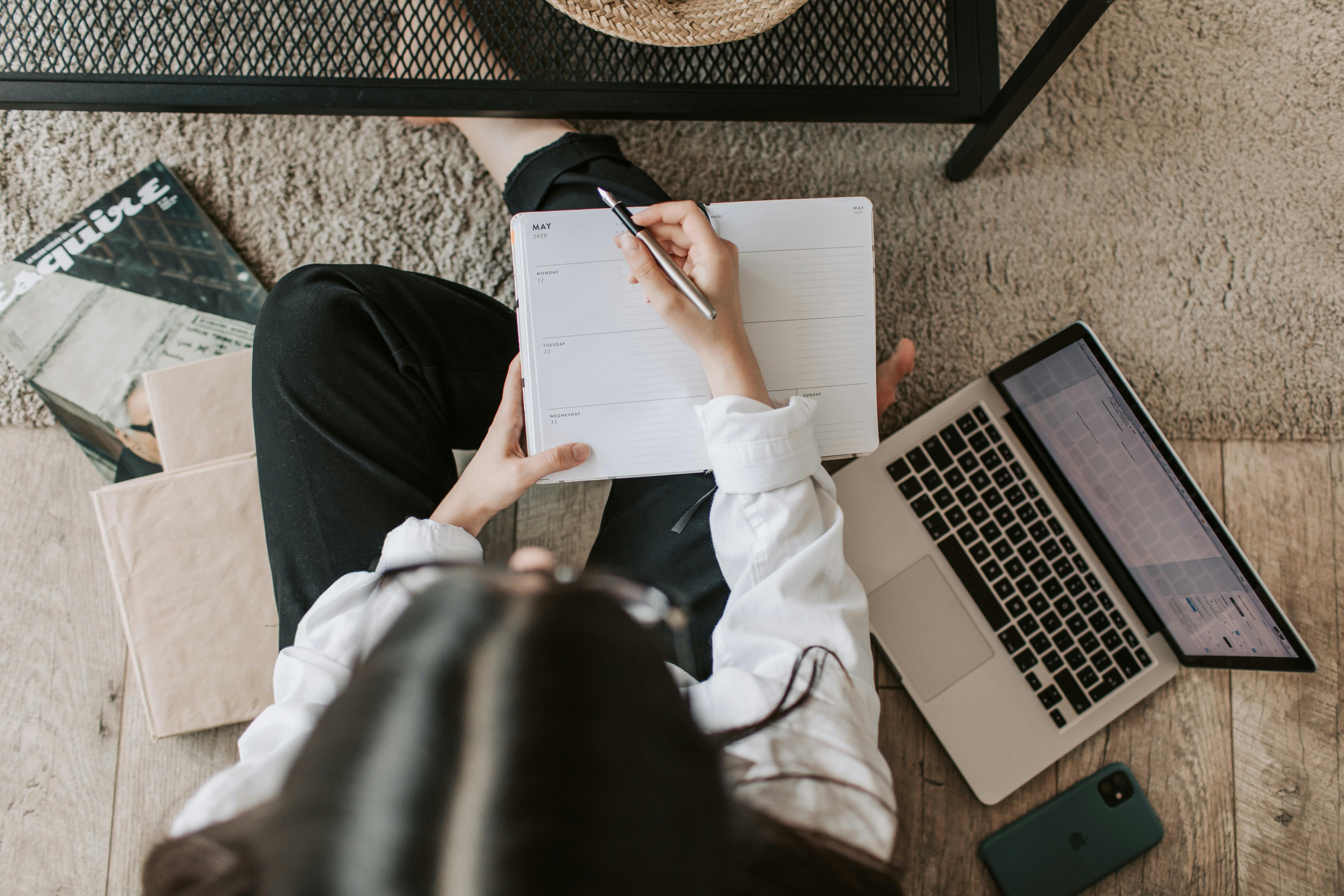 Woman sat on floor writing in diary with laptop next to her