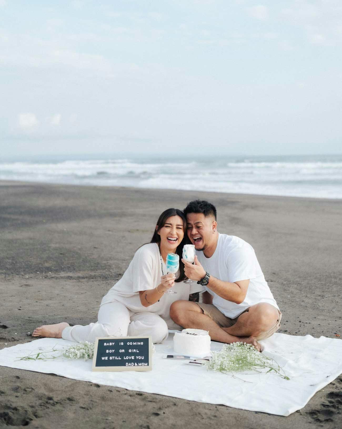 Couple laughing sitting on a beach holding cupcakes revealing the gender of their baby