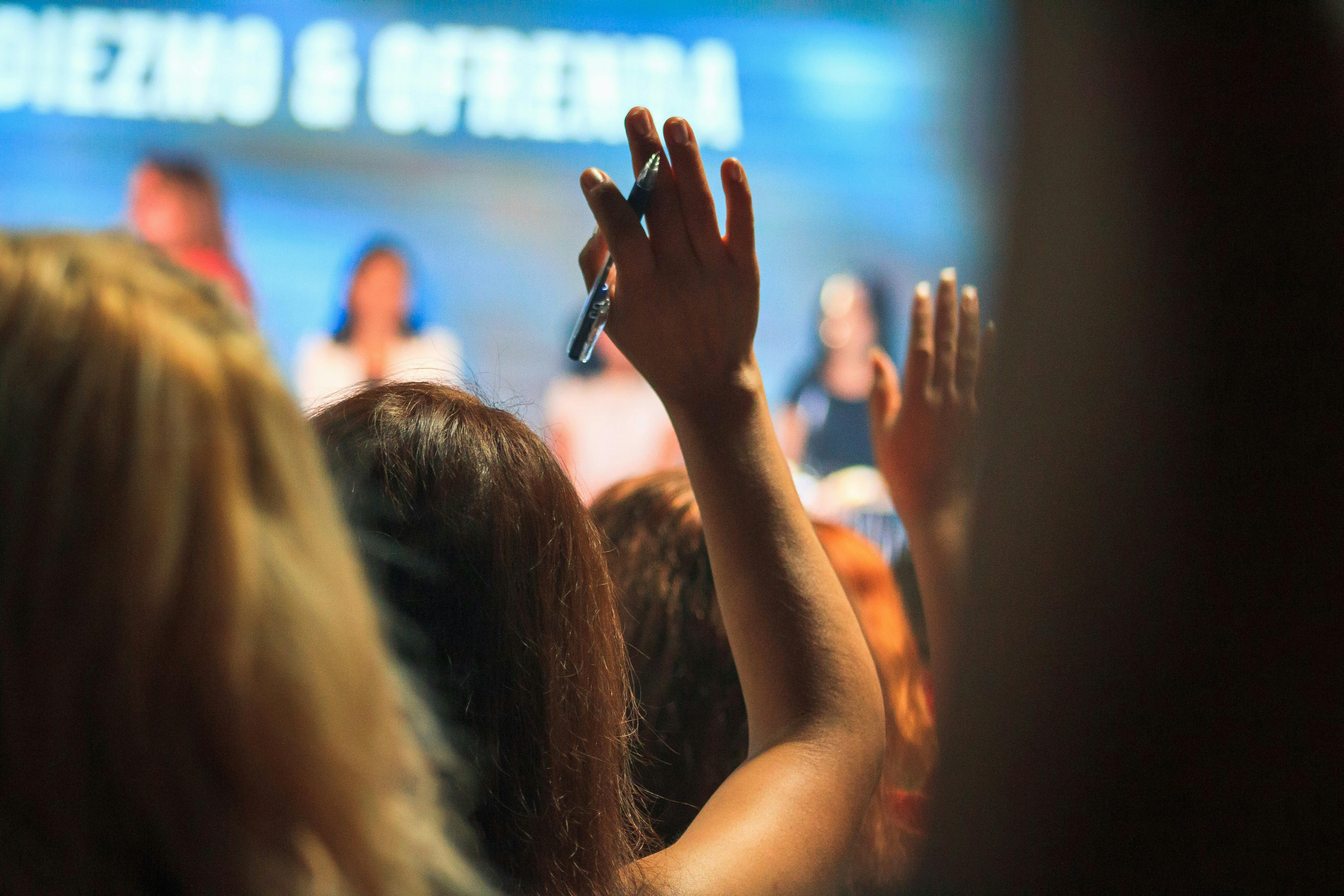 A close up of the back of people's heads in an audience for a conference event – some people with hands raised