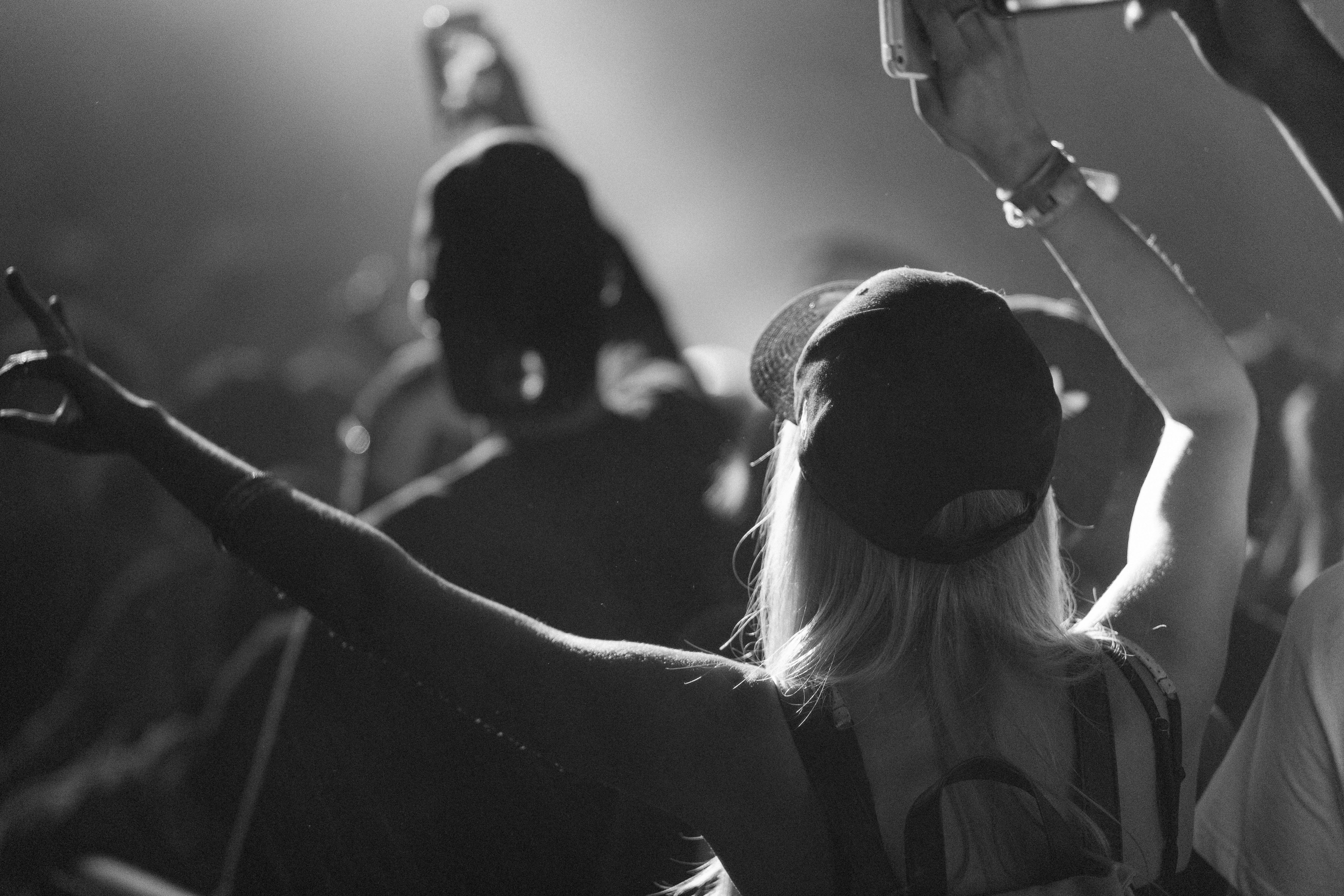 A black and white photo of an audience at a concert – focusing on back of woman with hands in air