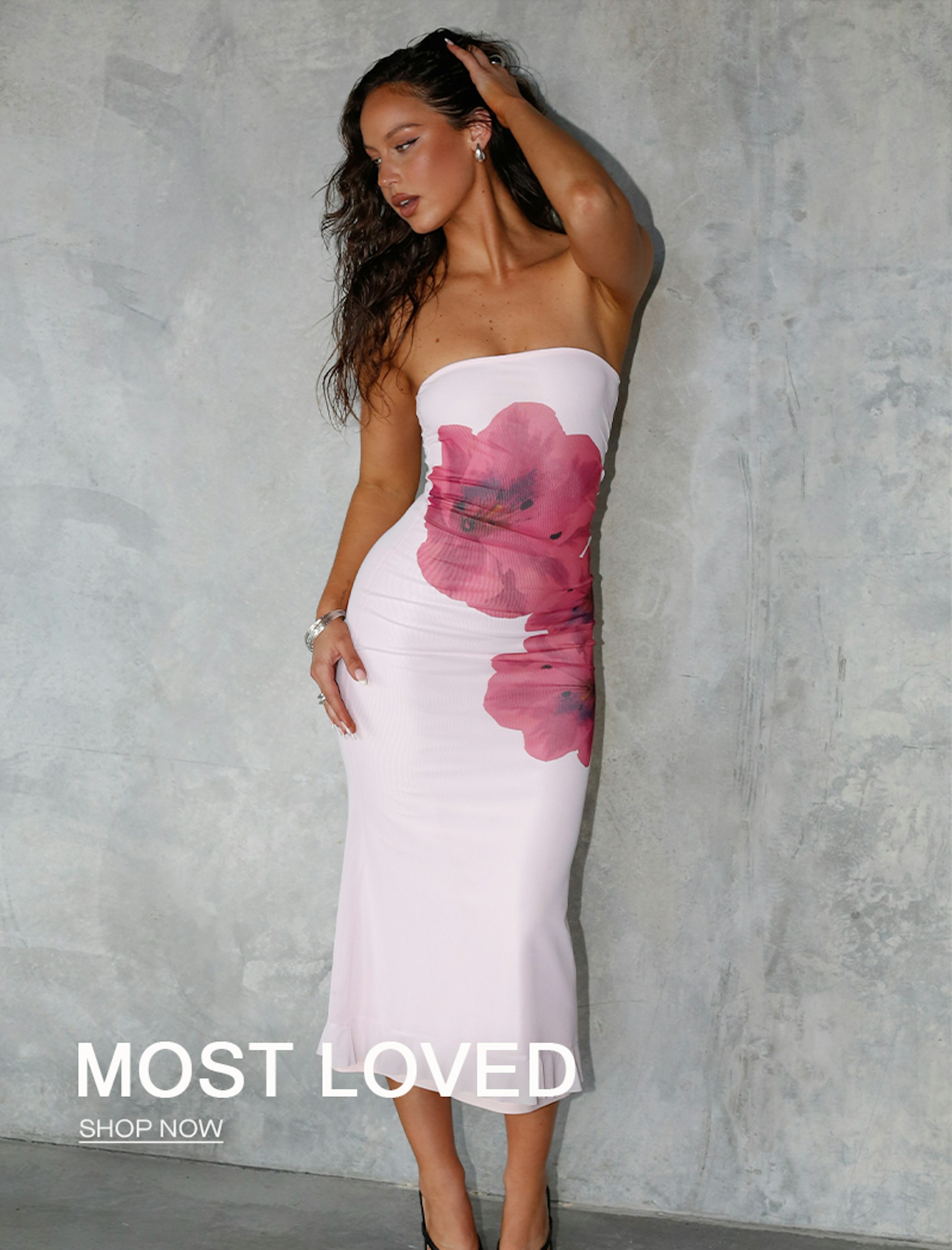 A woman with long hair is wearing a strapless white dress with large red flower prints. She poses against a concrete wall, touching her hair with one hand. Text over the image reads, "MOST LOVED. SHOP NOW.