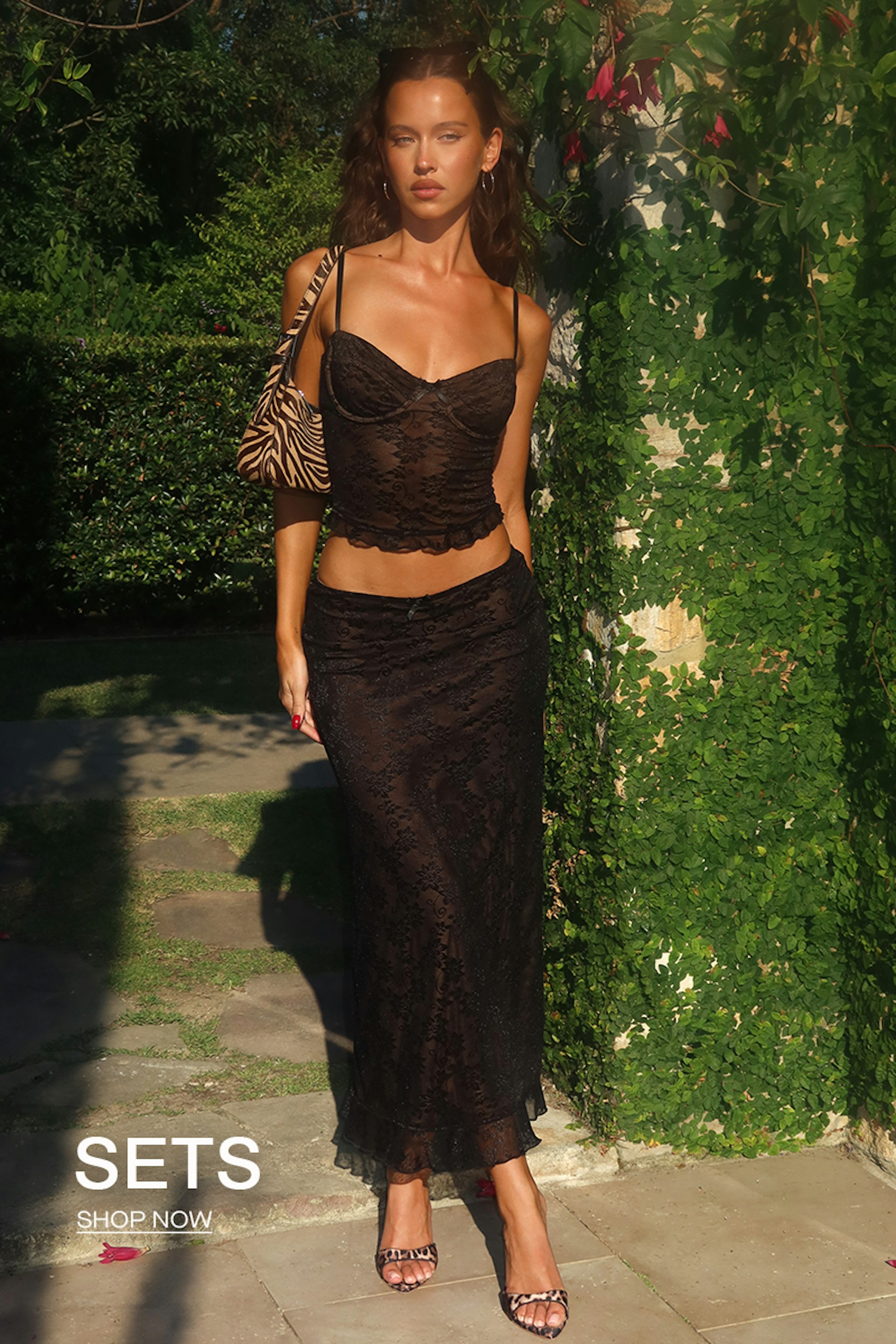 Woman in a black lace outfit stands outdoors next to greenery. She carries a zebra-patterned bag and wears strappy heels. Text in the bottom corner reads "SETS SHOP NOW." Sunlight creates soft shadows around her.