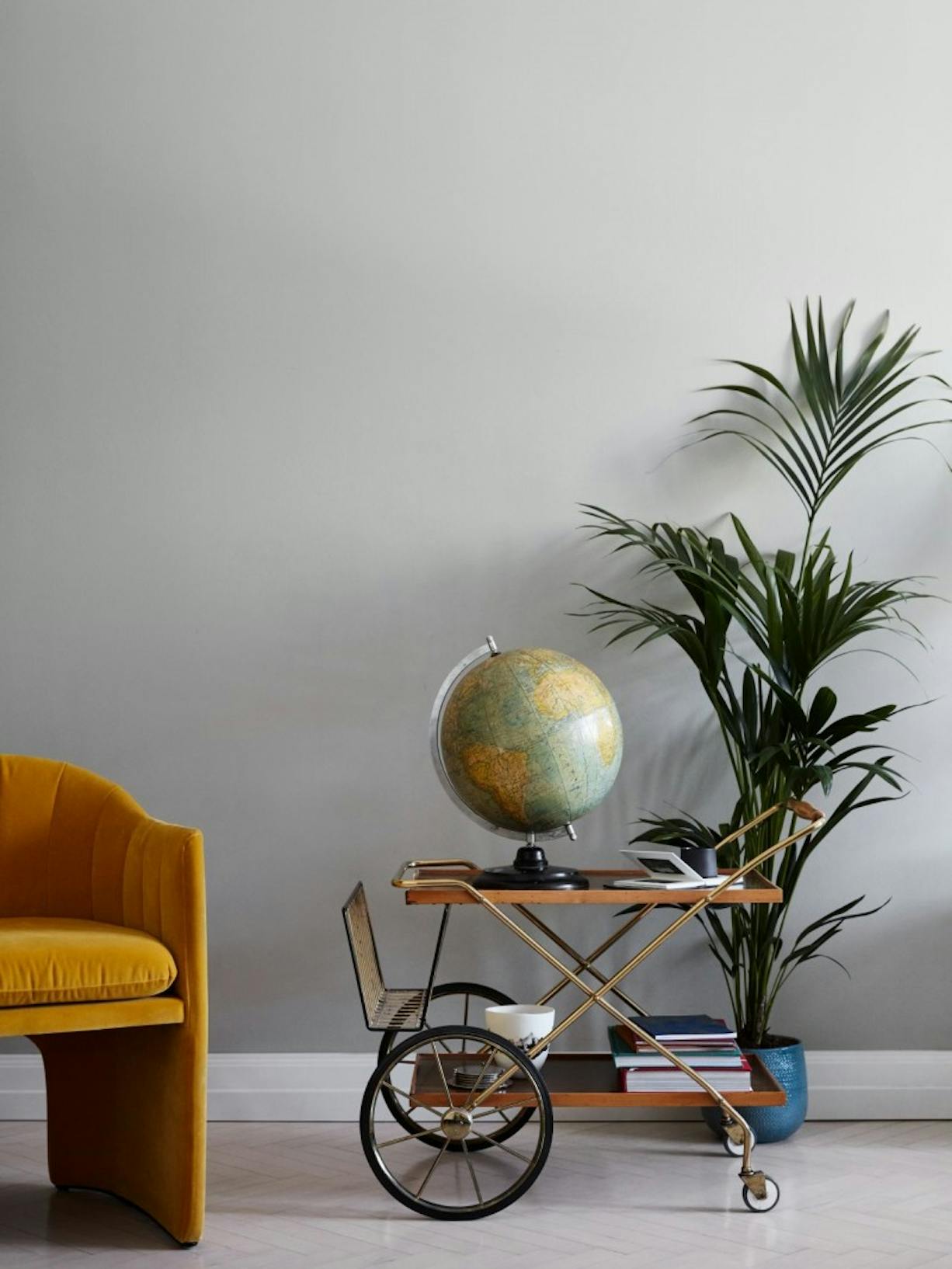 Yellow Armchair, Small Table with Black Wheels and Large Plant Against Grey 