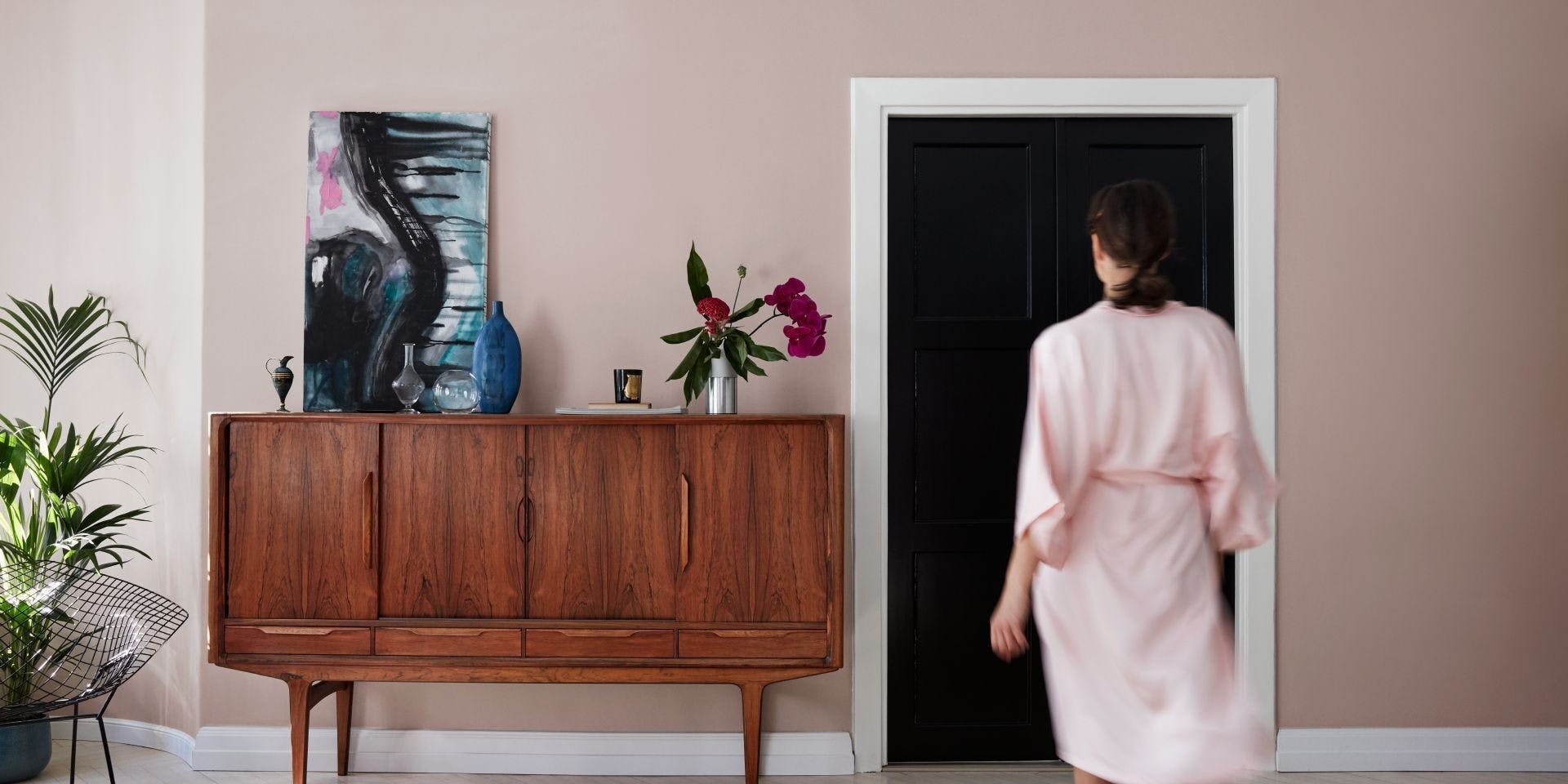 Back View of Woman in Pink Robe Walking Towards Door in Light Pink Room With Wooden Cabinet