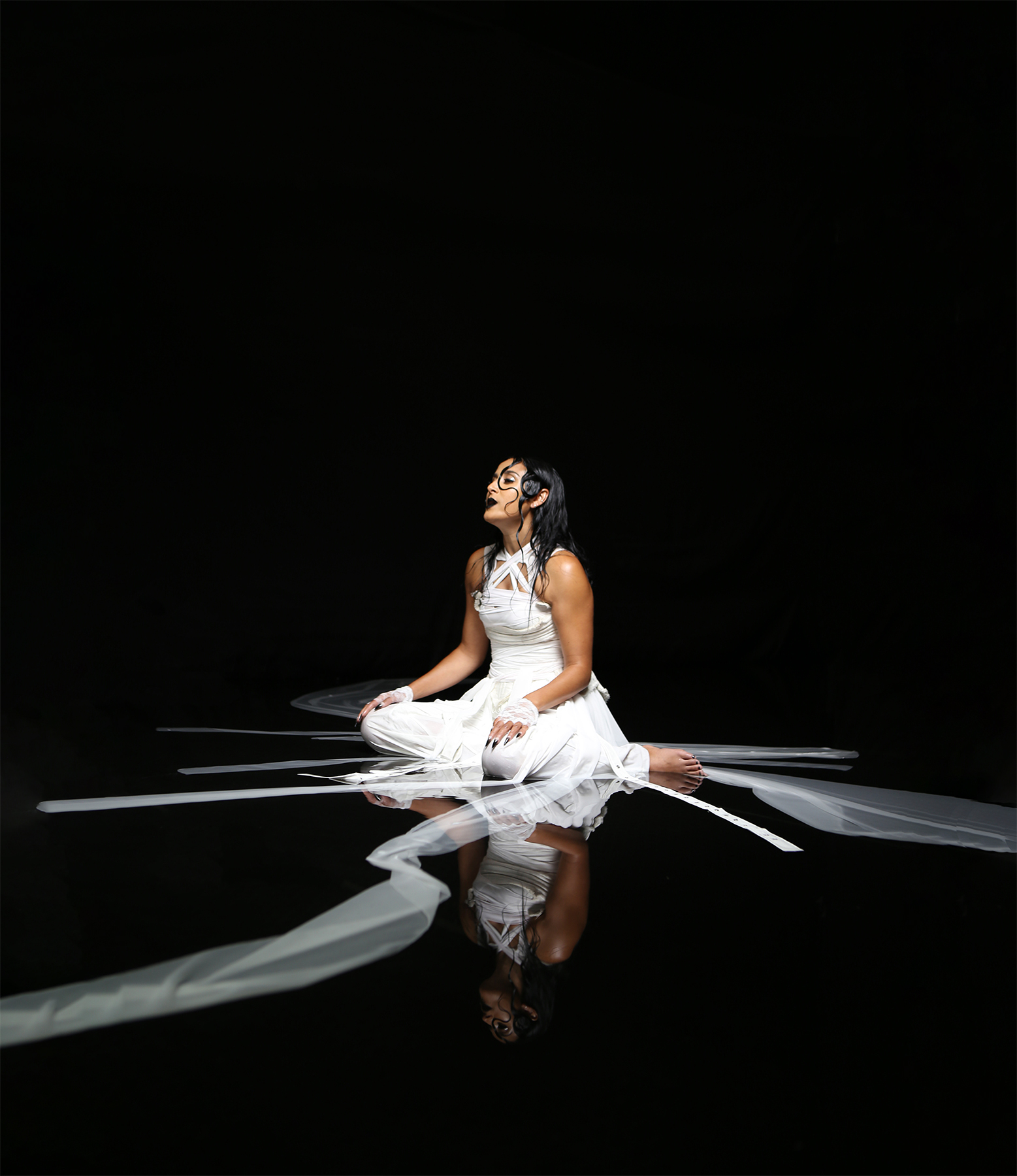 Fashion photography of a woman draped in long strips of white fabric in a black photography studio with water on the floor