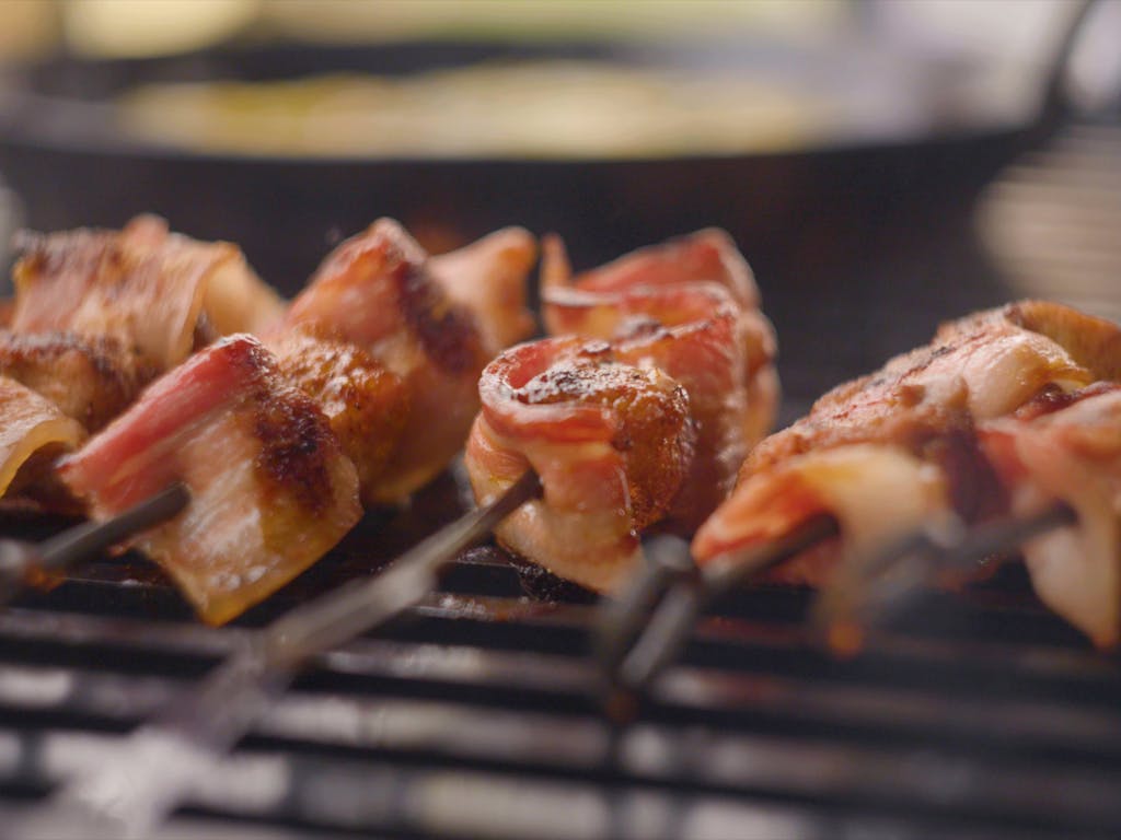 Crispy bacon and rabbit skewers cooking on a grill over fire