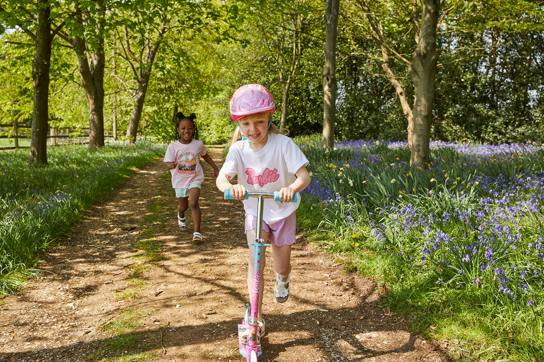 2 young girls playing in the woods, one runs, chasing the other, who is riding a scooter