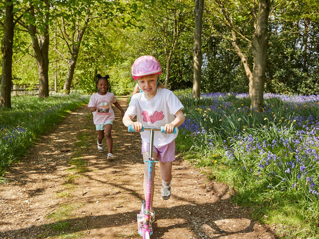 2 young girls playing in the woods, one runs, chasing the other, who is riding a scooter