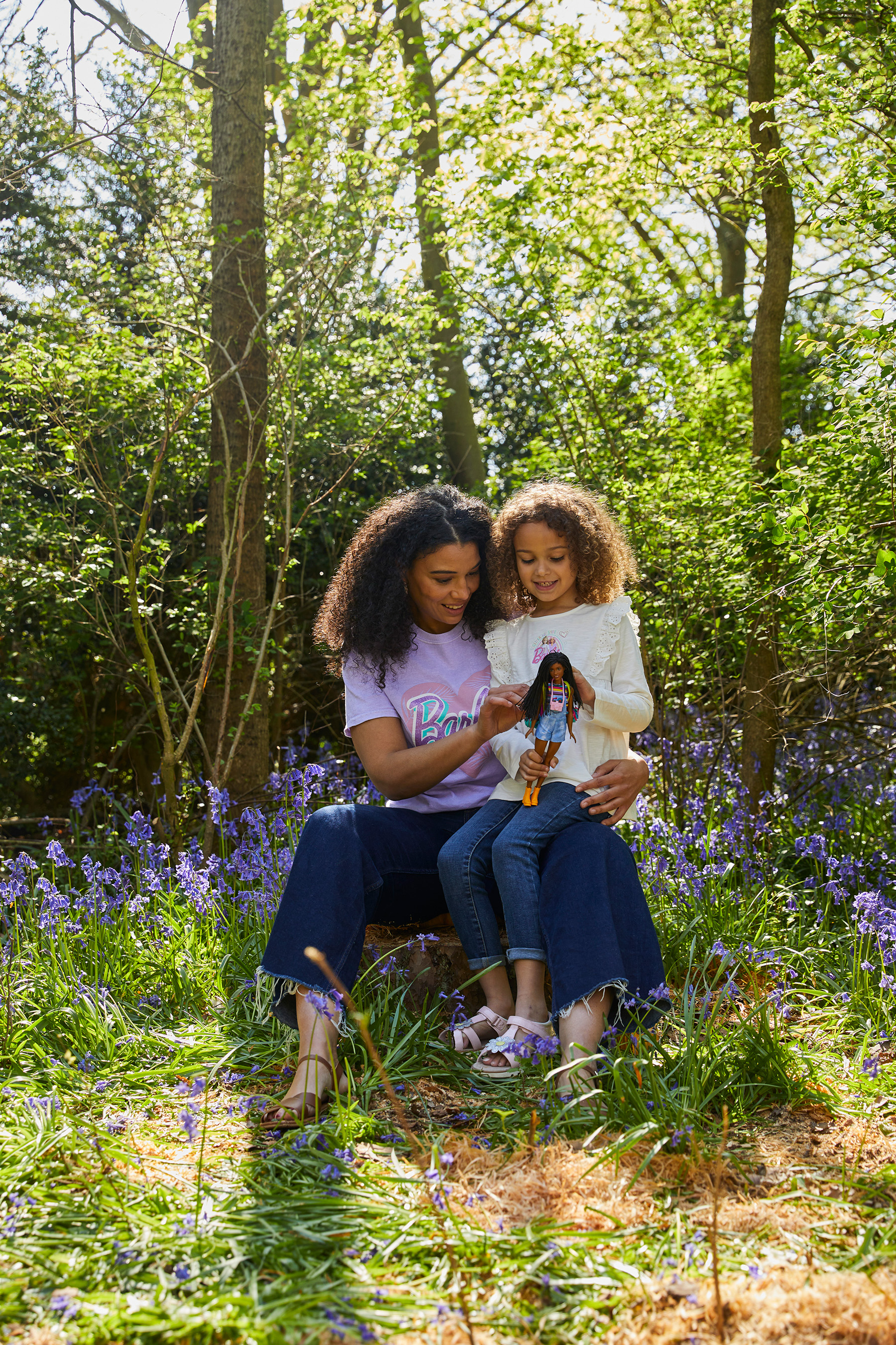 A woman and child, dressed in barbie tshirts, sit in a bluebell woodland, playing with a barbie doll