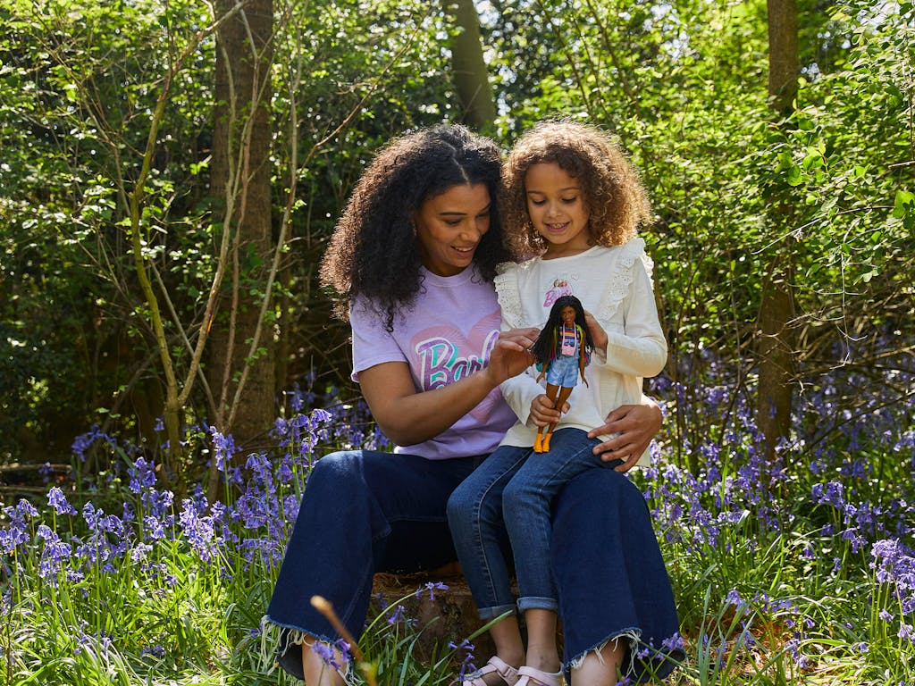 A woman and child, dressed in barbie tshirts, sit in a bluebell woodland, playing with a barbie doll