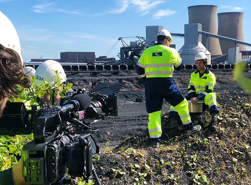 Two cameramen film staff in High-vis clothing, at a power station