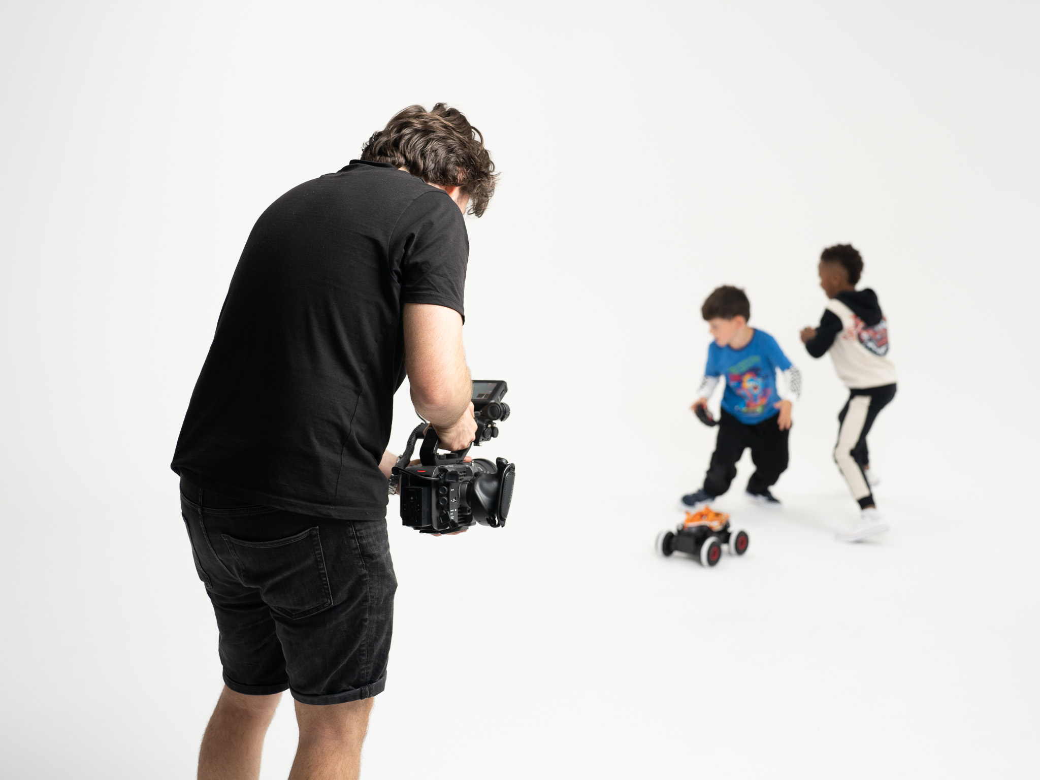 Cameraman films two young boys as they run around a white studio playing with a remote control car