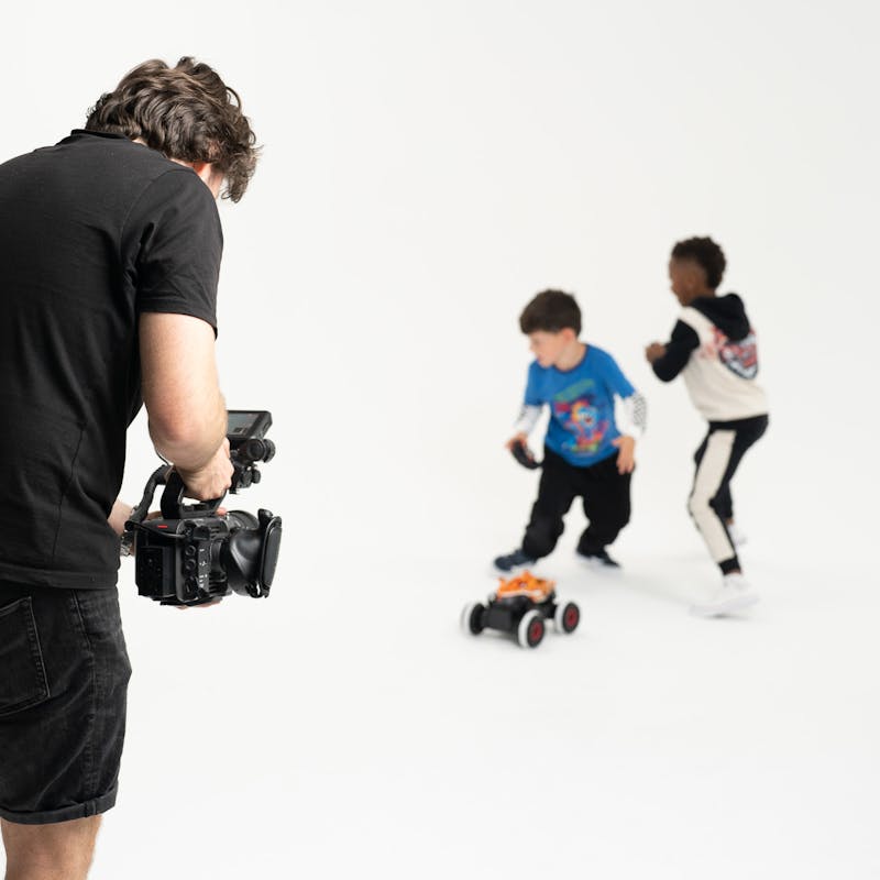Cameraman films two young boys as they run around a white studio playing with a remote control car