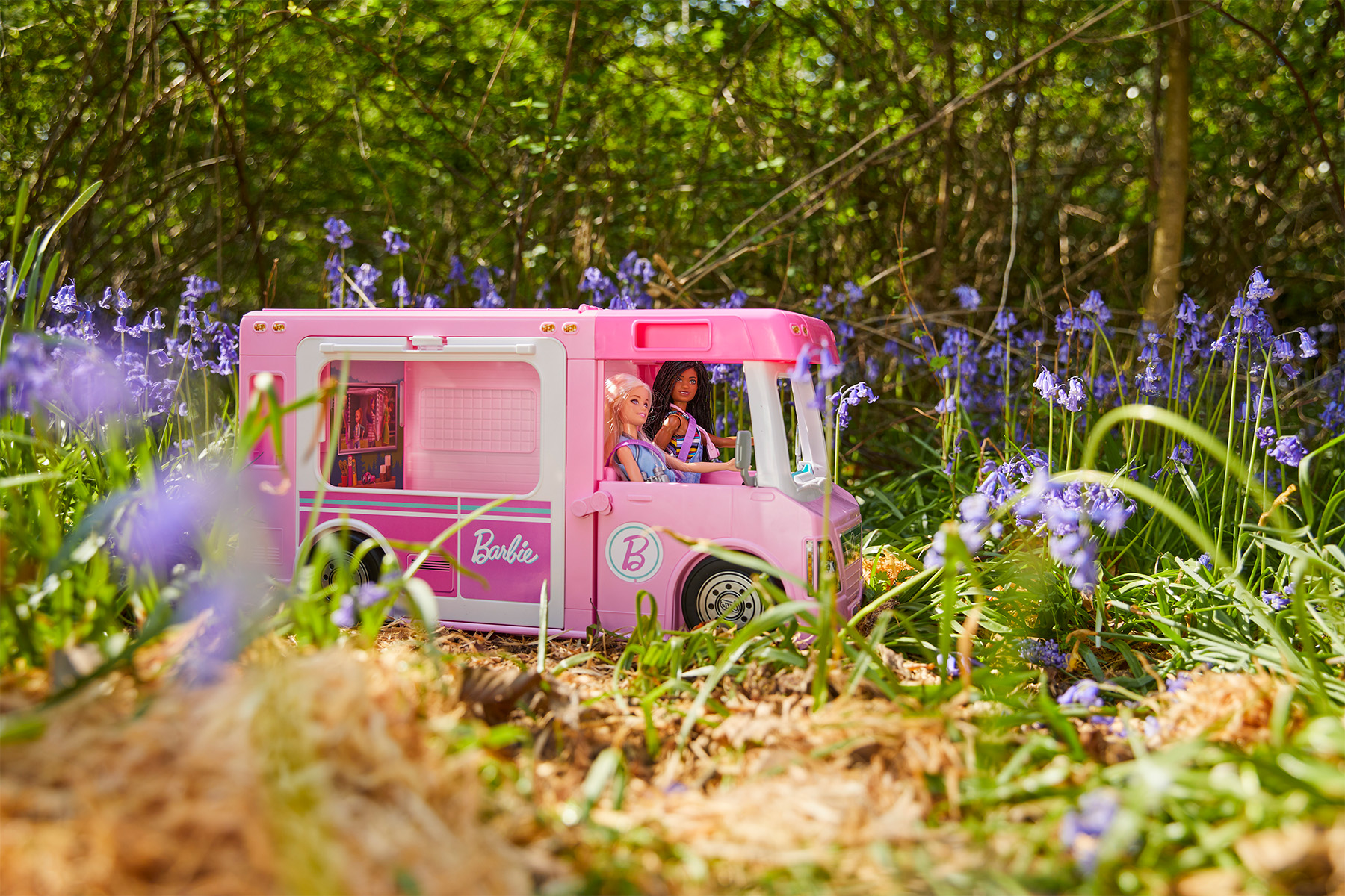 two barbie dolls driving a pink toy campervan in a bluebell woodland