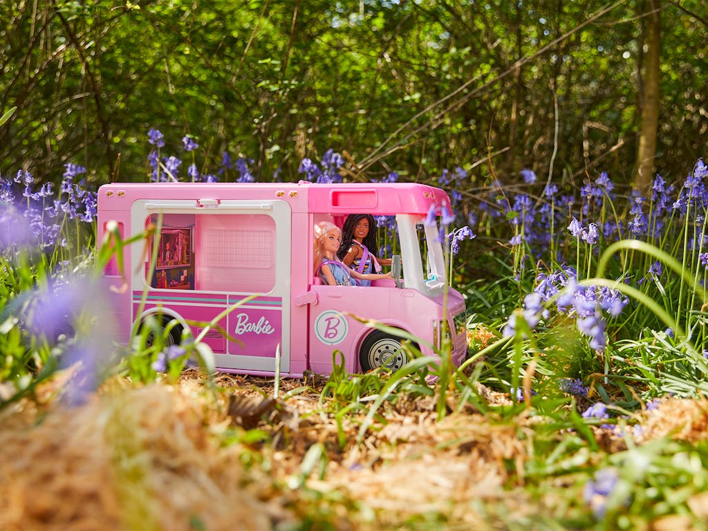 two barbie dolls driving a pink toy campervan in a bluebell woodland
