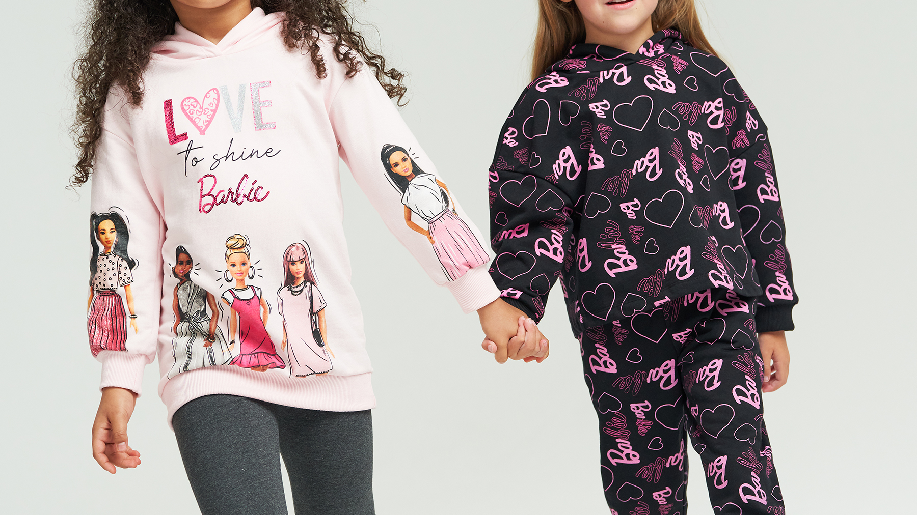 Two young girls, dressed in Barbie tracksuits walk forwards through a white photography studio holding hands 