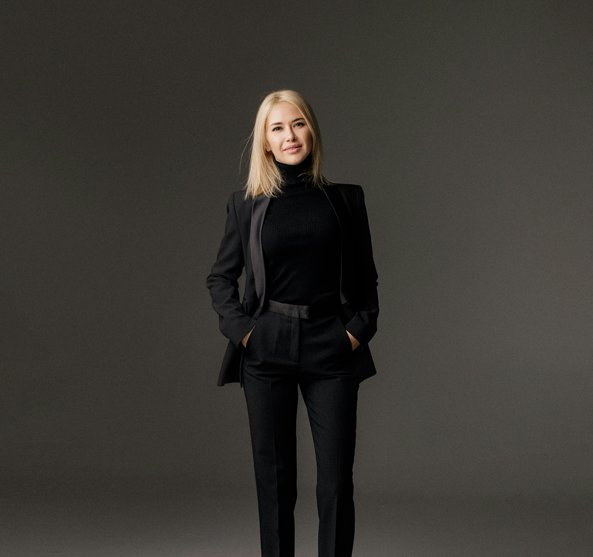 A trendy blonde woman in black jeans and a black suit jacket, poses for a photo, smiling with hands in her pockets
