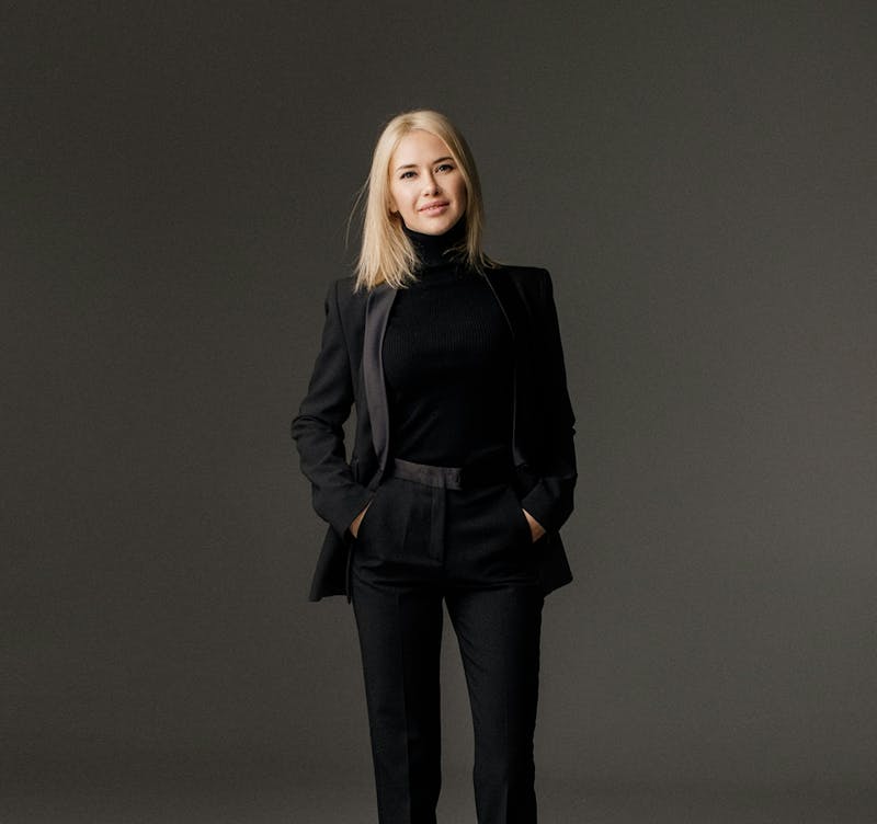 A trendy blonde woman in black jeans and a black suit jacket, poses for a photo, smiling with hands in her pockets