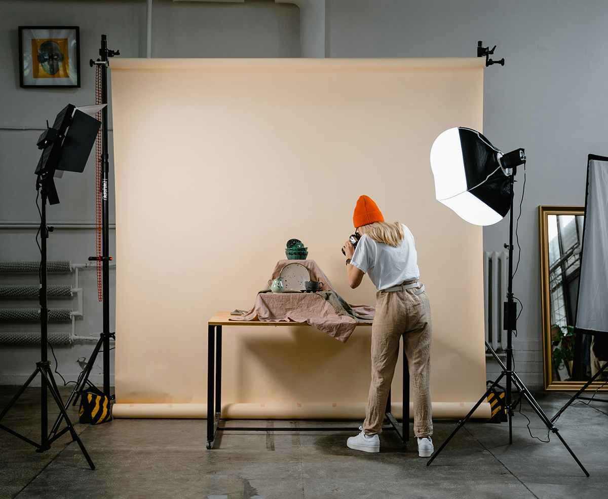 A woman wearing an orange beanie hat, takes photographs of ceramics products, in front of peach coloured photography background roll