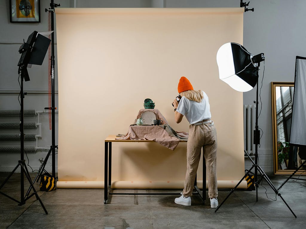 A woman wearing an orange beanie hat, takes photographs of ceramics products, in front of peach coloured photography background roll
