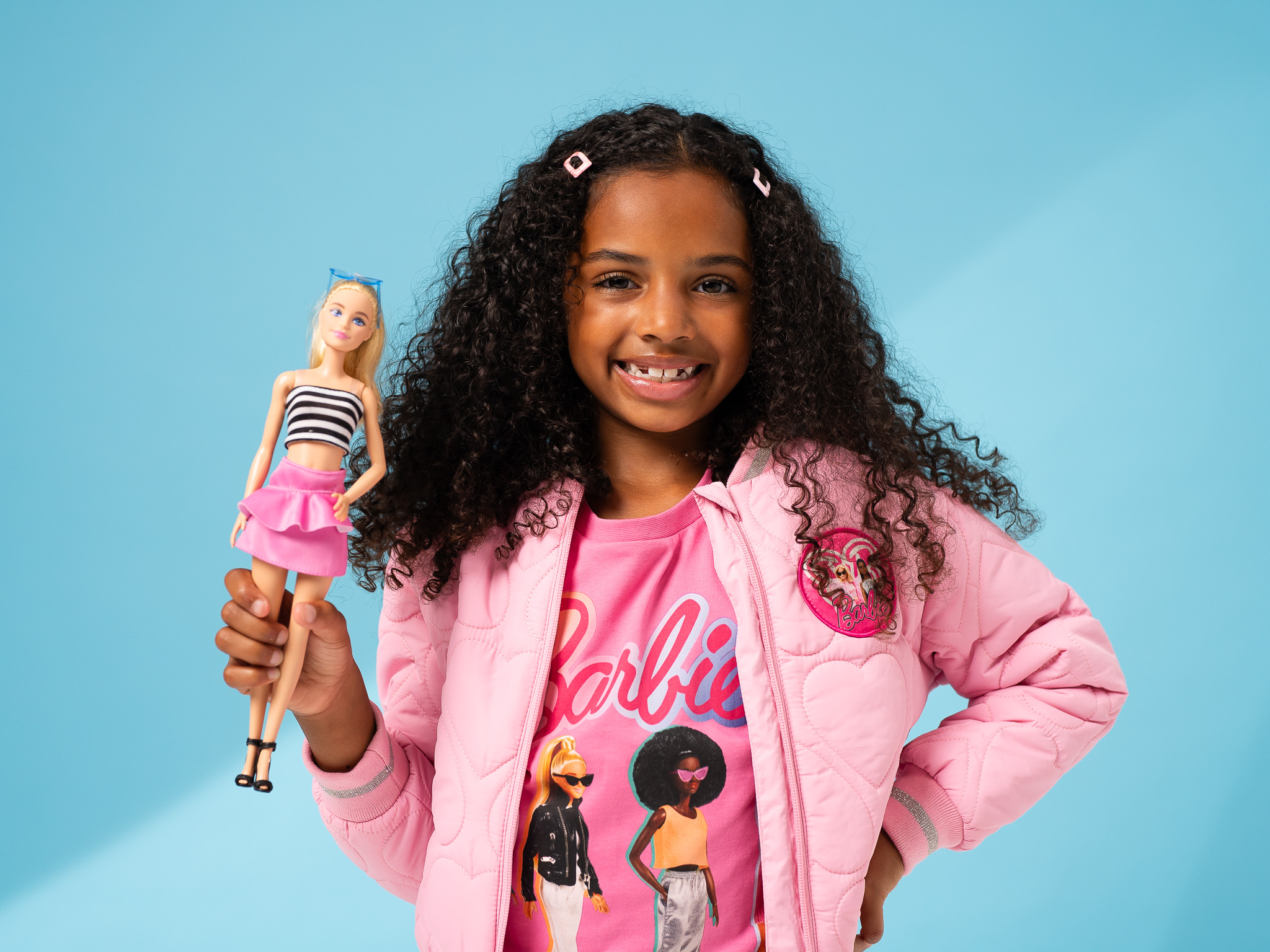 child poses with hand on hip, holding a barbie doll, wearing a pink barbie jacket