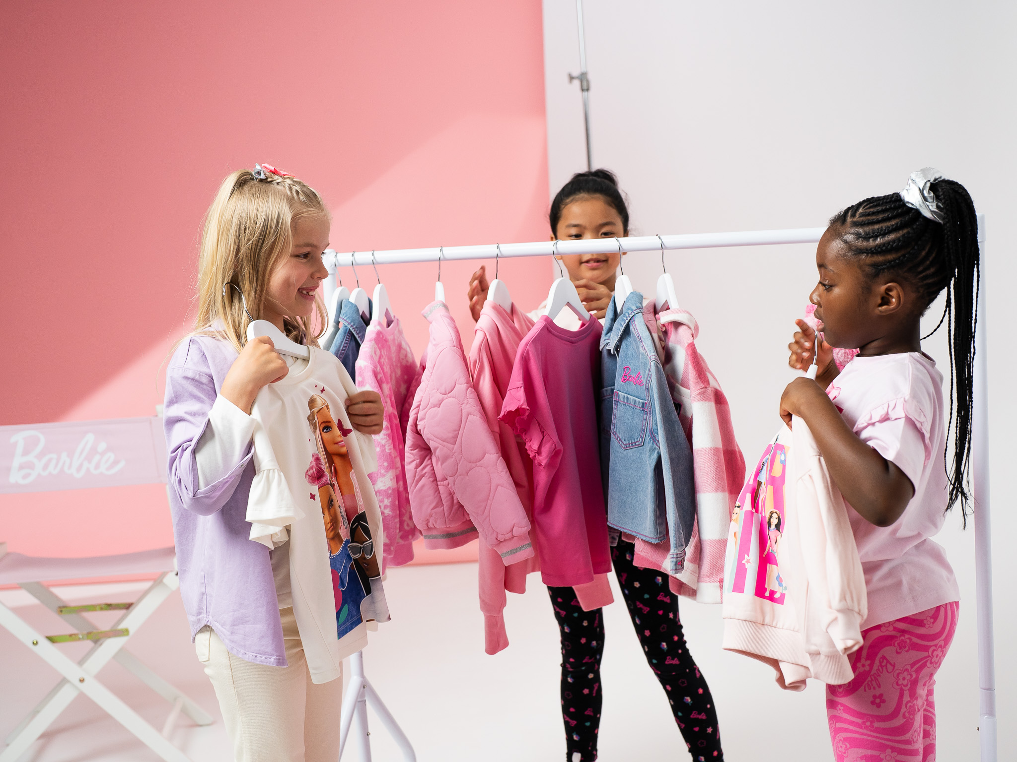3 girls standing next to a clothing rail, hold up barbie outfits on hangers