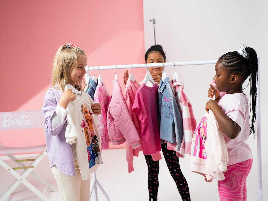 3 girls standing next to a clothing rail, hold up barbie outfits on hangers