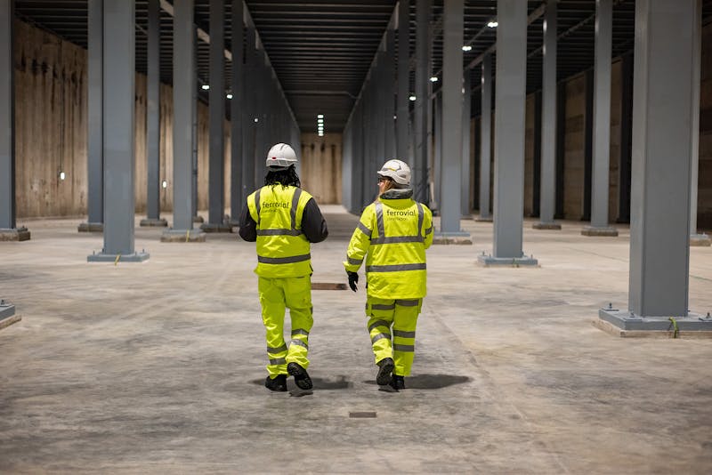 Two colleagues, dressed in high-vis, walk through large industrial hanger, filled with metal structural pillars