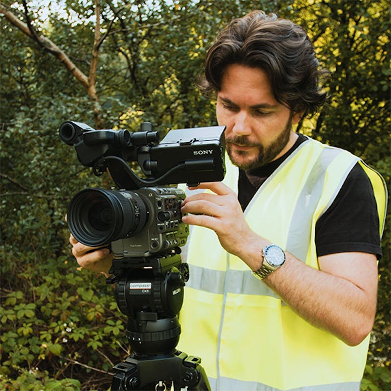 Cameraman in high-vis filming setting up video camera with green trees behind
