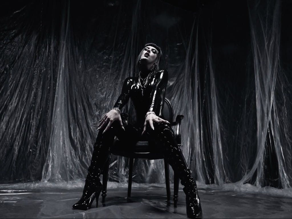 Woman dressed in black latex sat in a chair in a room lined with plastic sheets